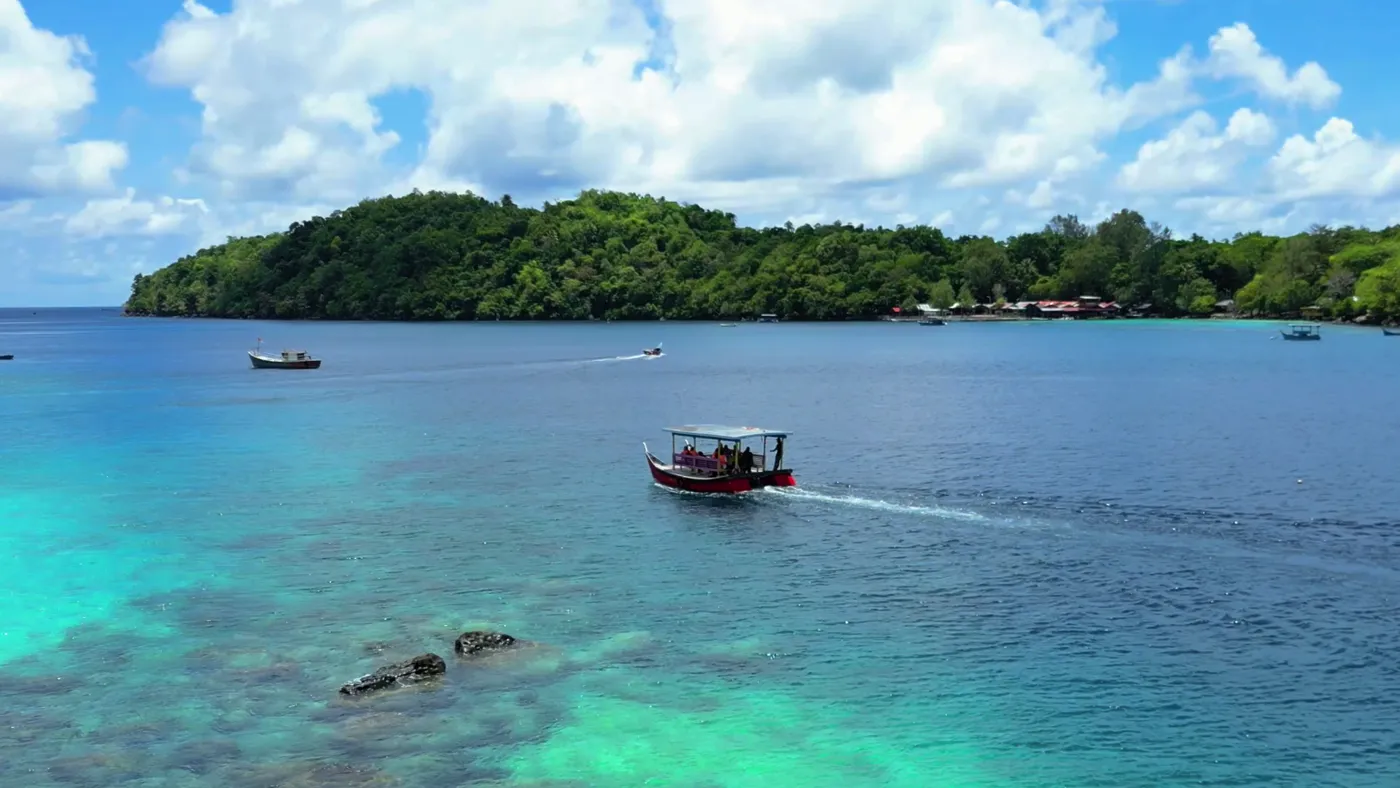 A boat crossing clear turquoise water over a reef, with a forested island behind, off Pulau Weh